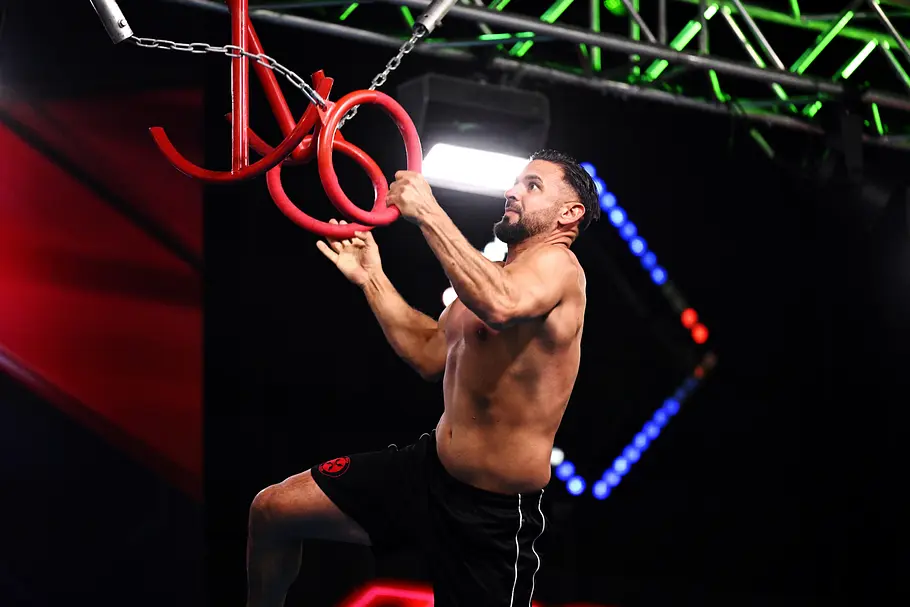 Ali Moussa ist Teilnehmer bei Ninja Warrior Germany 2026 Ali Moussa hält sich mit nacktem Oberkörper und schwarzer Shorts an roten Ringen eines beleuchteten Hindernisparcours fest.