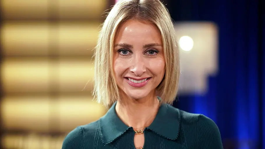 Alina Bock lächelt mit blondem Bob und dunkelgrünem Oberteil vor einem unscharfen, gelb und blau beleuchteten Hintergrund.