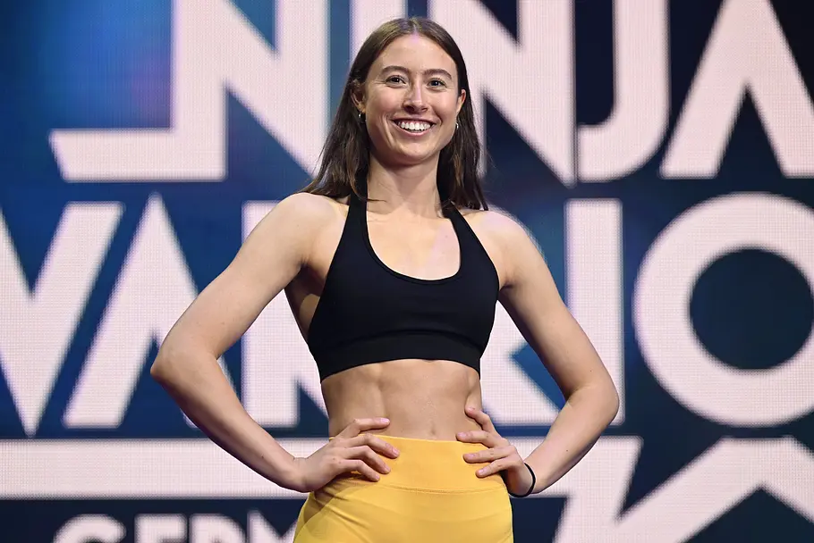 Alina Tornau ist Teilnehmer bei Ninja Warrior Germany 2026 Alina Tornau steht lächelnd im schwarzen Sporttop und gelber Hose mit den Händen an den Hüften vor einer Schriftwand.