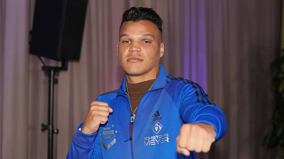 Allen Bauer Allen Bauer posiert in einer blauen Trainingsjacke mit erhobenen Fäusten vor einem Vorhang. Eine Faust streckt er nach vorn.