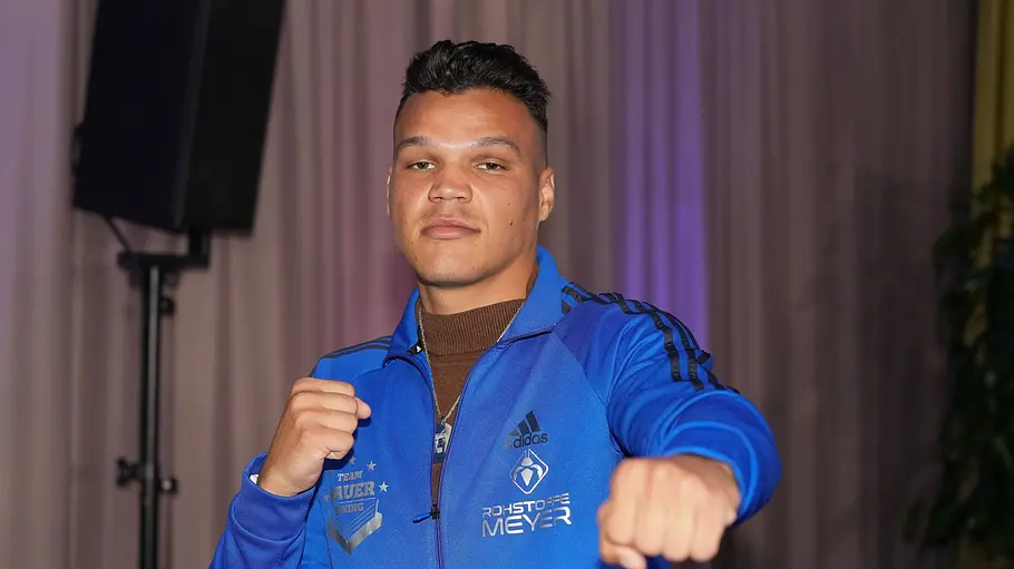 Allen Bauer Fame Fighting Allen Bauer posiert in blauer Trainingsjacke mit erhobenen Fäusten vor einem Vorhang. Eine Faust streckt er zur Kamera.