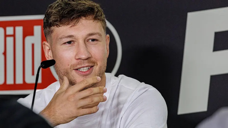 Oliver Ginkel Fame Fighting Oliver Ginkel sitzt im weißen T-Shirt vor einem Mikrofon, eine Hand am Kinn. Hinter ihm ist ein Bild-Logo zu sehen.
