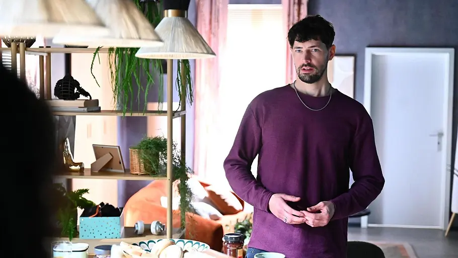 Jan steht verlegen in Imanis Wohnung. Er trägt einen Bordeauxroten Pulli