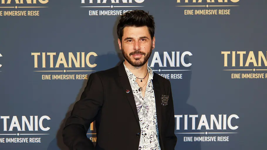 Riccardo Angelini auf dem Red Carpet der Titanic-Ausstellung. Er trägt einen Anzug und ein Hemd mit einigen offenen Knöpfen.