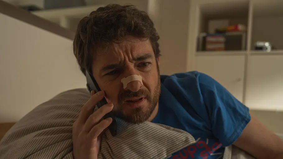 Alpha Males Gorka Otxoa liegt mit einem Telefon in der Hand in einem Bett. Er hat ein Pflaster über der Nase.