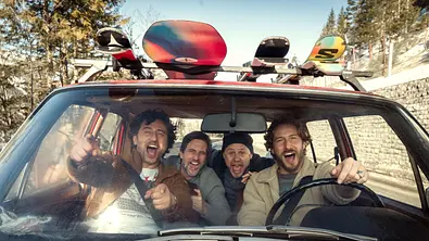 Vier Männer sitzen fröhlich lachend in einem Auto und schauen aus der Frontscheibe. Auf dem Dach des roten Autos sind bunte Snowboards befestigt, im Hintergrund sieht man verschneite Berge und Wald. - Foto: © 2025 Netflix, Inc.