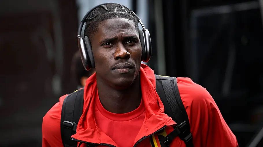 Ein belgischer Nationalspieler in roter Teamkleidung mit Kopfhörern und Rucksack kommt vor einem Länderspiel am Stadion an. Sein Blick ist nach vorne gerichtet, im Hintergrund ist der Mannschaftsbus zu sehen.