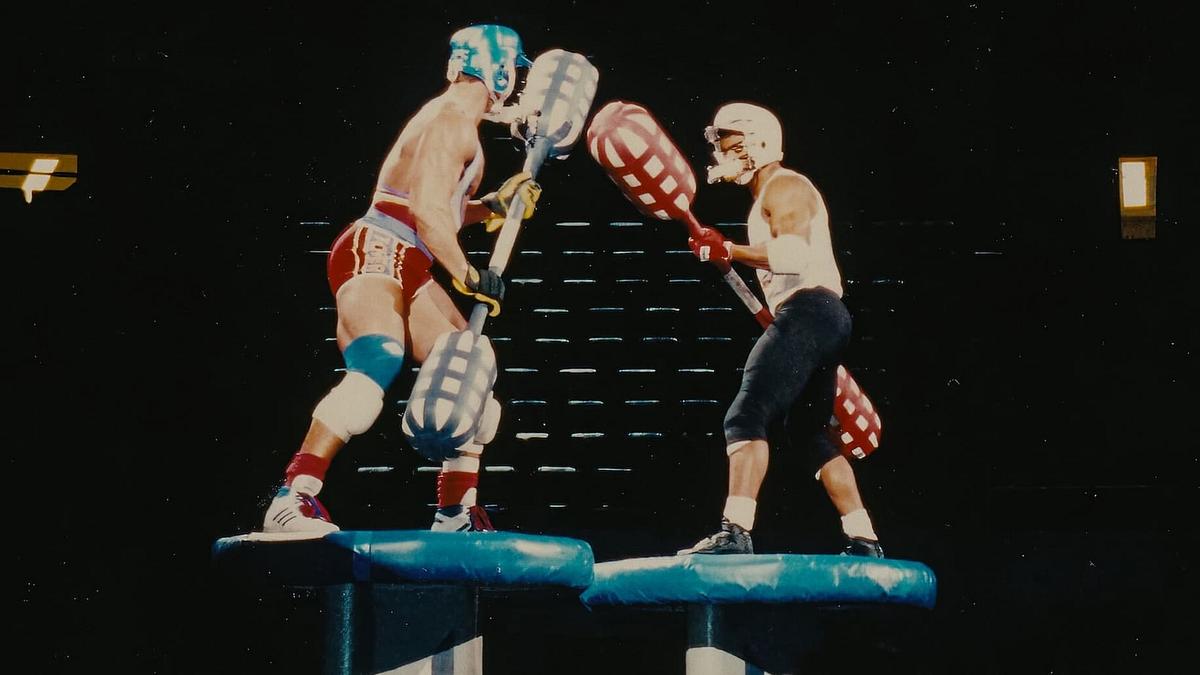 American Gladiators kehrt zurück. Zwei Teilnehmer der Sendung American Gladiators stehen sich auf zwei Plattformen gegenüber und haben lange Kampfstäbe in ihren Händen.