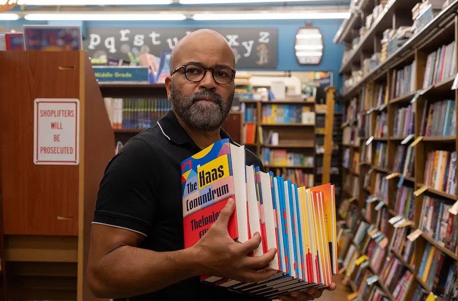 Thelonious „Monk“ Ellison (Jeffrey Wright) steht mit Brille und einem Stapel bunter Bücher zwischen Bücherregalen.