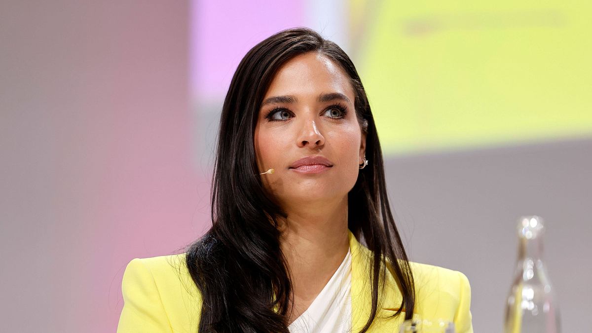 Amira Aly als Moderatorin mit offenen Haaren, ernster Mine und in einem gelben Blazer gekleidet. 