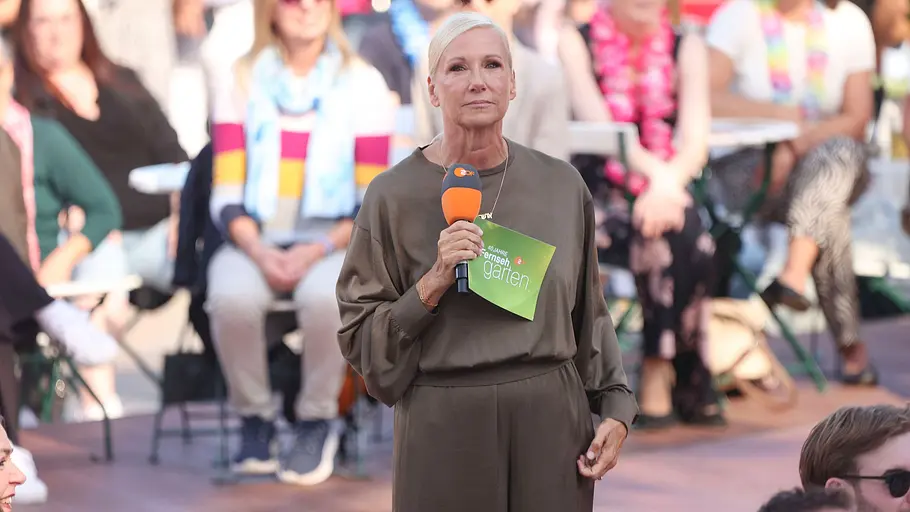 Andrea Kiewel hat ihr nächstes Projekt schon gefunden. Andrea Kiewel steht in einem olivgrauen Kleid mit Mikrofon und grüner Moderationskarte vor einem sitzenden Publikum.