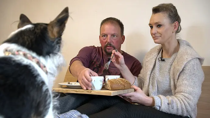 Ein Mann und eine Frau sitzen gemeinsam auf einer Sitzbank und frühstücken. Die Frau hält ein Holzbrett mit zwei Tassen und einem Brot in der Hand, während der Mann nach einer Tasse greift. Im Vordergrund sitzt ein schwarz-weißer Hund mit dem Rücken zur Kamera und blickt die beiden an. Die Szene wirkt gemütlich und spielt sich in einem schlicht eingerichteten Innenraum ab. - Foto: RTL Ein Mann und eine Frau sitzen gemeinsam auf einer Sitzbank und frühstücken. Die Frau hält ein Holzbrett mit zwei Tassen und einem Brot in der Hand, während der Mann nach einer Tasse greift. Im Vordergrund sitzt ein schwarz-weißer Hund mit dem Rücken zur Kamera und blickt die beiden an. Die Szene wirkt gemütlich und spielt sich in einem schlicht eingerichteten Innenraum ab. - Foto: RTL