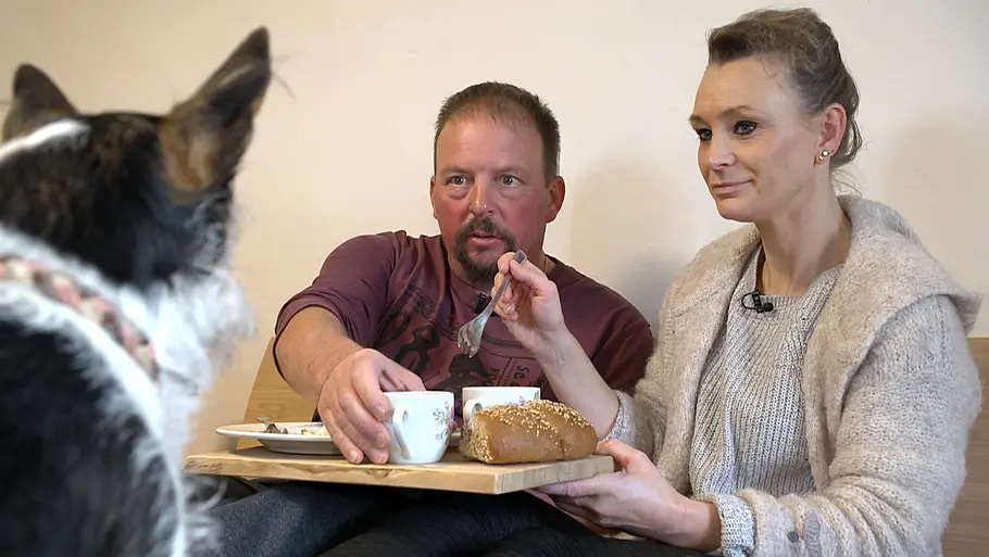 Andreas und Katerina Ein Mann und eine Frau sitzen gemeinsam auf einer Sitzbank und frühstücken. Die Frau hält ein Holzbrett mit zwei Tassen und einem Brot in der Hand, während der Mann nach einer Tasse greift. Im Vordergrund sitzt ein schwarz-weißer Hund mit dem Rücken zur Kamera und blickt die beiden an. Die Szene wirkt gemütlich und spielt sich in einem schlicht eingerichteten Innenraum ab.