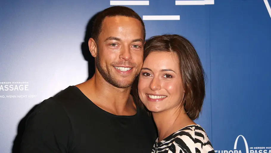 Andrej Mangold und Jennifer Lange Andrej Mangold im schwarzen Shirt und Jennifer Lange im Zebramuster-Oberteil stehen lächelnd vor einer blauen Werbewand.