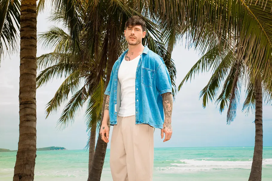 Angelo Carlucci Angelo Carlucci steht am Strand unter Palmen vor türkisfarbenem Meer und trägt ein offenes blaues Hemd über weißem Shirt sowie helle, weite Hose.