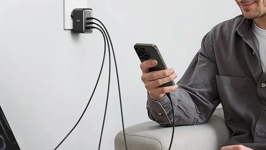 Anker Adapter Ein Mehrfach-USB-C-Ladegerät von Anker steckt in einer Wandsteckdose und versorgt über drei Kabel mehrere Geräte. Eine Person sitzt auf einem Sofa und hält ein Smartphone, das mit dem Ladegerät verbunden ist.