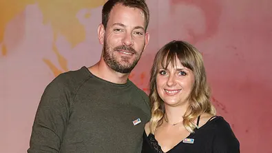 Anna und Gerald stehen Arm in Arm vor einer wolkigen Wand. Beide lächeln.  - Foto: IMAGO / BREUEL-BILD