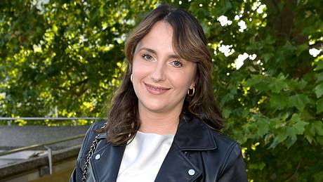 Anna Heiser steht vor ein paar Bäumen in einer Halbtotalen und lächelt leicht. Sie trägt eine schwarze Lederjacke und zwei Ohrringe.  - Foto: IMAGO / Future Image
