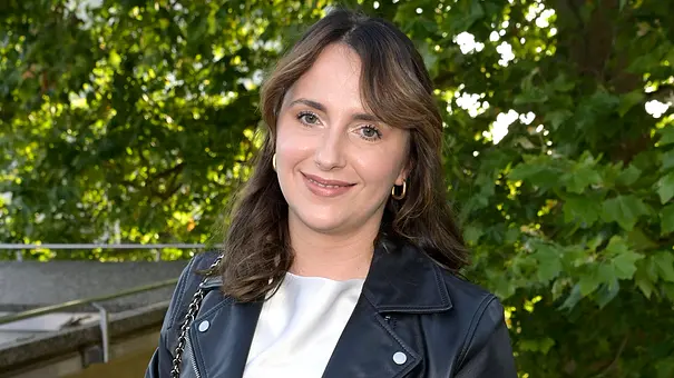 Anna Heiser steht vor ein paar Bäumen in einer Halbtotalen und lächelt leicht. Sie trägt eine schwarze Lederjacke und zwei Ohrringe.  - Foto: IMAGO / Future Image