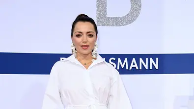 Anne Menden posiert in einem weißen Hemdkleid auf dem roten Teppich und hält eine Hand auf ihrem Babybauch - Foto: IMAGO / Future Image