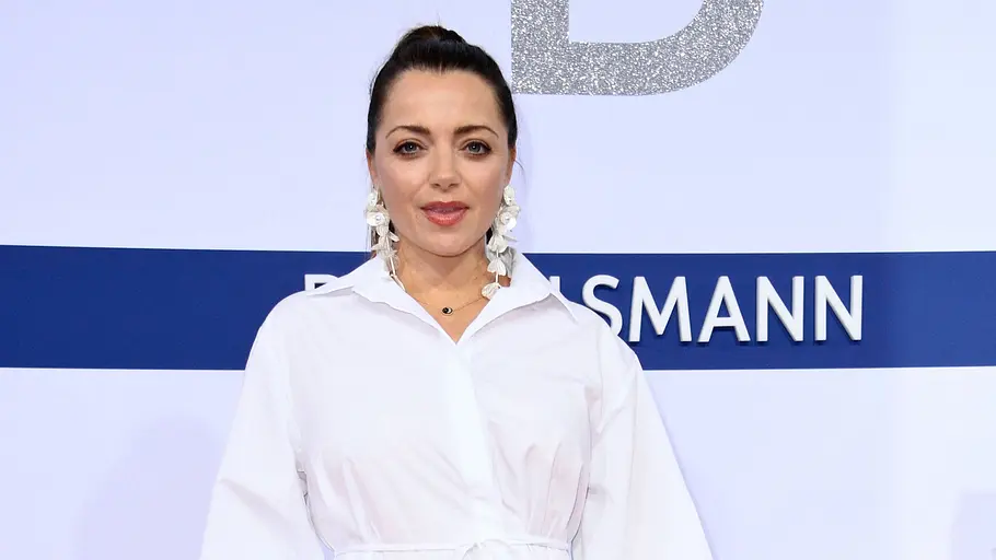 Anne Menden Anne Menden posiert in einem weißen Hemdkleid auf dem roten Teppich und hält eine Hand auf ihrem Babybauch