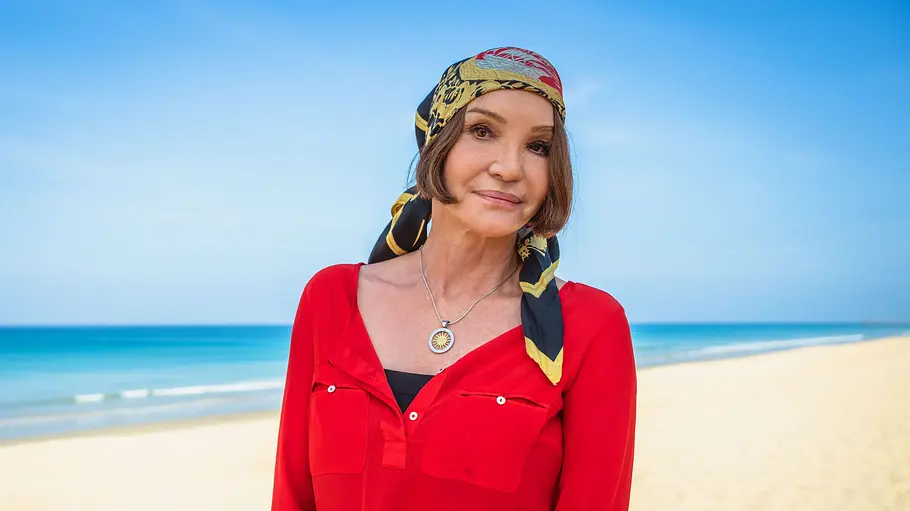 Anouschka Renzi war schon in einigen Reality-TV-Formaten zu sehen Anouschka Renzi steht in roter Bluse und gemustertem Kopftuch an einem Sandstrand vor türkisblauem Meer.