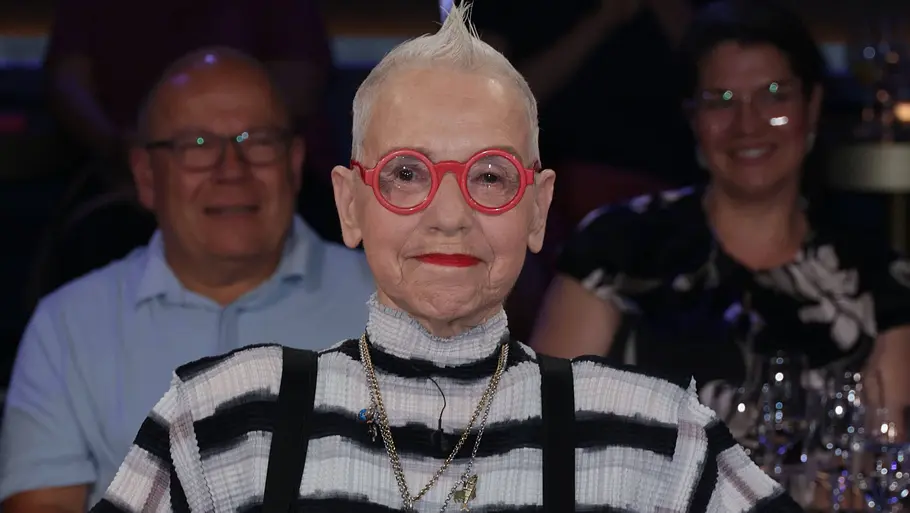 Antje Vogel Antje Vogel sitzt in einem Sessel im Studio einer Talkshow. Sie trägt ein schwarz‑weiß gestreiftes Oberteil mit schwarzen Hosenträgern, mehrere Ketten und Ringe. Im Vordergrund stehen Wassergläser und Blumen, im Hintergrund ist das Publikum unscharf zu sehen.