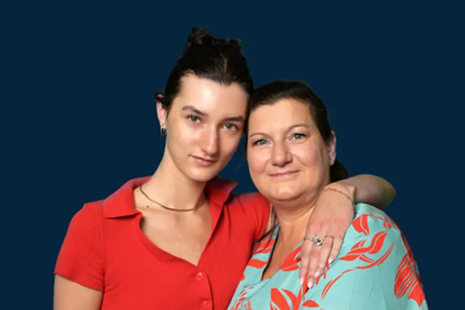 Anya Elsner in rotem Oberteil legt den Arm um ihre Mutter Katrin in türkis-roter Bluse. Beide stehen vor dunkelblauem Hintergrund.