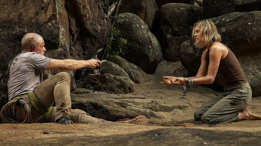 Szene aus dem Film Apex auf Netflix: Ben (Taron Egerton) sitzt verletzt in der Schlucht und bedroht Sasha (Charlize Theron) aus der Ferne mit einem Messer.