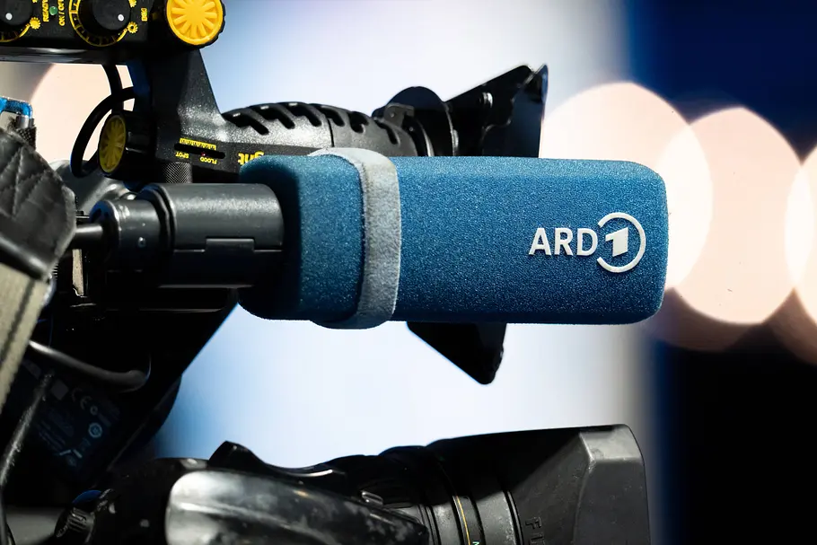 Ein Mikrofon mit blauem Windschutz und weißem ARD-Logo ist an einer Fernsehkamera befestigt. Im Hintergrund leuchten unscharfe Lichter.