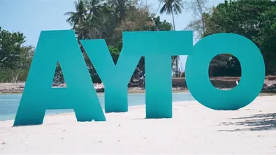 Große türkisfarbene Buchstaben bilden „AYTO“ auf einem Sandstrand. Dahinter liegen Wasser und Bäume mit Palmen. - Foto: RTL