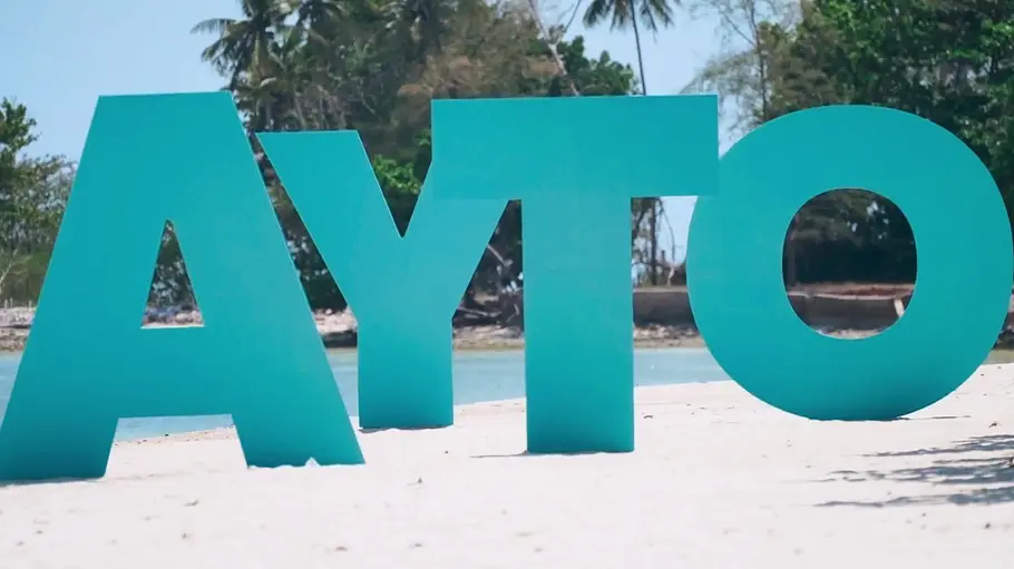 AYTO Große türkisfarbene Buchstaben bilden „AYTO“ auf einem Sandstrand. Dahinter liegen Wasser und Bäume mit Palmen.