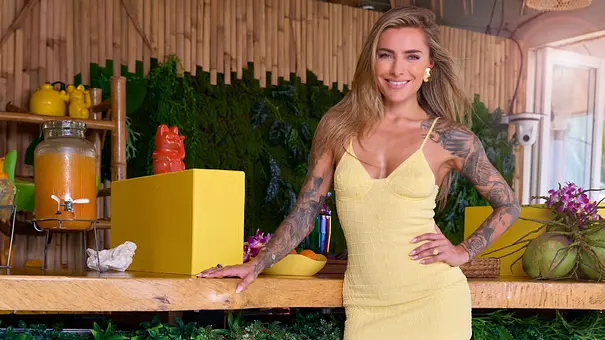 Reality-TV-Moderatorin Sophia Thomalla steht lächelnd in einem gelben Sommerkleid an einer dekorierten Bar mit Pflanzen und tropischem Ambiente. - Foto: RTL / Frank Beer