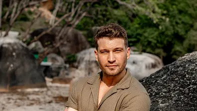 Raul Richter steht mit verschränkten Armen an einem großen Felsen an einem Strand, im Hintergrund sind Bäume und blauer Himmel zu sehen. - Foto: RTL / Katya Bulgakova