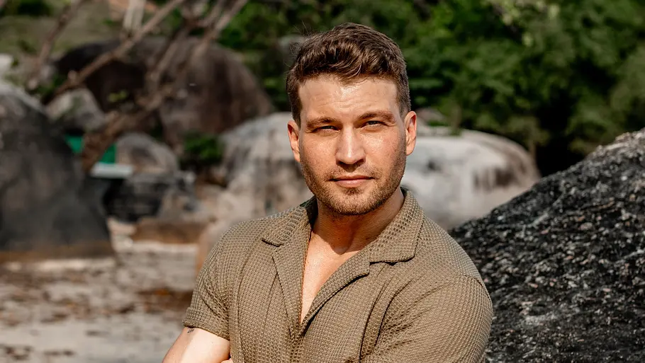 Raul Richter Raul Richter steht mit verschränkten Armen an einem großen Felsen an einem Strand, im Hintergrund sind Bäume und blauer Himmel zu sehen.
