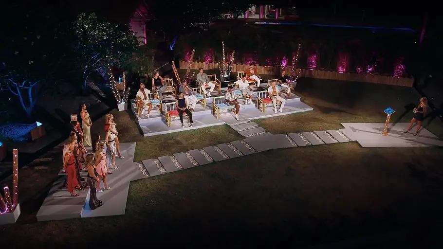 AYTO Nachtszene einer Matching Night: Kandidatinnen und Kandidaten stehen und sitzen auf beleuchteten Podesten im Garten, rechts steht eine Moderatorin auf einer kleinen Bühne.