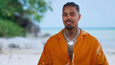 Ein Mann mit geflochtenen Haaren, Tattoos und Silberketten trägt ein offenes orangefarbenes Hemd vor einer Strandkulisse. - Foto: RTL