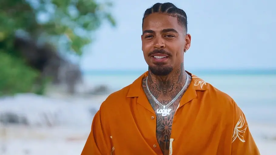Germain Ein Mann mit geflochtenen Haaren, Tattoos und Silberketten trägt ein offenes orangefarbenes Hemd vor einer Strandkulisse.