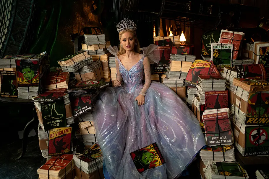Ariana Grande als Glinda in Wicked Teil 2 Ariana Grande als Glinda sitzt inmitten von Plakaten über die Böse Hexe des Westens. Sie trägt eine Krone und ein außergwöhnliches Kleid. Sie schaut verängstigt.