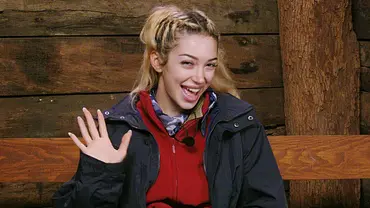 Ariel mit blonden Haaren sitzt im Dschungelcamp auf einer Holzbank, trägt Outdoor-Kleidung und winkt lachend in die Kamera. Im Hintergrund sind Holzwände und ein Baumstamm zu sehen. - Foto: RTL Ariel mit blonden Haaren sitzt im Dschungelcamp auf einer Holzbank, trägt Outdoor-Kleidung und winkt lachend in die Kamera. Im Hintergrund sind Holzwände und ein Baumstamm zu sehen. - Foto: RTL