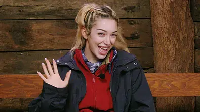 Ariel mit blonden Haaren sitzt im Dschungelcamp auf einer Holzbank, trägt Outdoor-Kleidung und winkt lachend in die Kamera. Im Hintergrund sind Holzwände und ein Baumstamm zu sehen. - Foto: RTL
