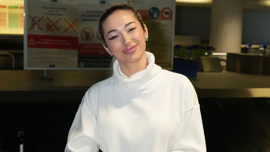 Ariel Hediger Ariel Hediger trägt einen weißen Pullover und lächelt in die Kamera. Sie hat ihre Hände auf zwei vor ihr stehenden Koffern gelegt und steht vor einem Gepäckband am Flughafen.