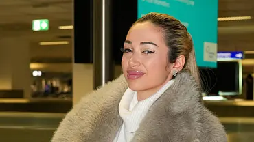Ariel sitzt an einem Flughafen auf einem Gepäckband, trägt einen hellen Rollkragenpullover und eine flauschige Jacke. - Foto: IMAGO / BOBO Ariel sitzt an einem Flughafen auf einem Gepäckband, trägt einen hellen Rollkragenpullover und eine flauschige Jacke. - Foto: IMAGO / BOBO