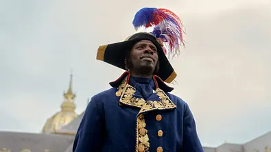 Assane Diop trägt eine blaue Uniform mit Goldstickerei und einen Hut mit rot-weiß-blauen Federn vor einem Kuppelgebäude. - Foto: Emmanuel Guimier/Netflix Assane Diop trägt eine blaue Uniform mit Goldstickerei und einen Hut mit rot-weiß-blauen Federn vor einem Kuppelgebäude. - Foto: Emmanuel Guimier/Netflix