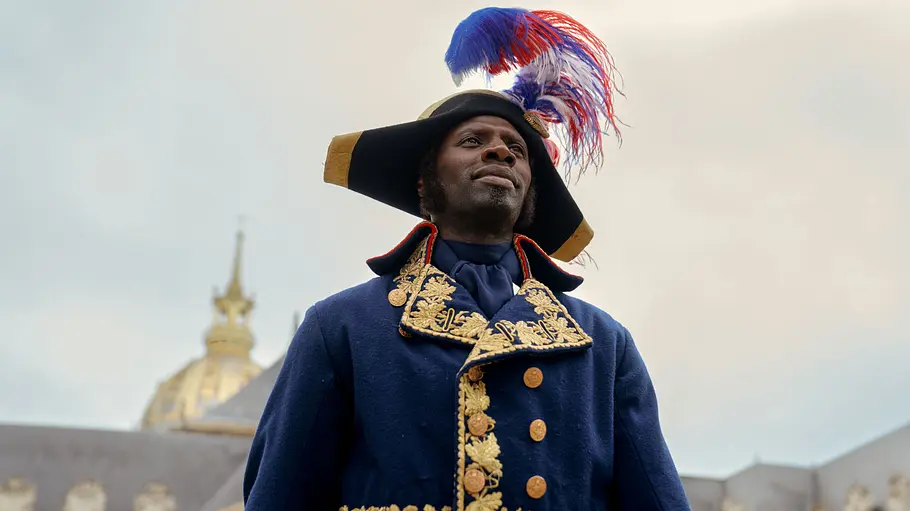 Assane Diop ist ein Meister der Täuschung, doch seine neue Stimme ist leider keiner seiner Tricks. Assane Diop trägt eine blaue Uniform mit Goldstickerei und einen Hut mit rot-weiß-blauen Federn vor einem Kuppelgebäude.