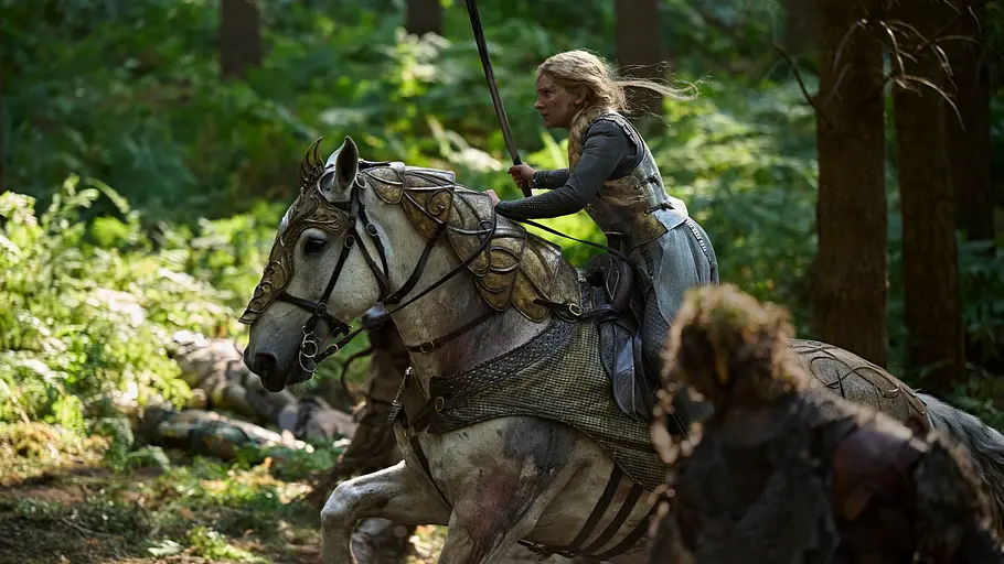 Galadriel reitet in Rüstung mit erhobenem Schwert auf einem gepanzerten weißen Pferd durch den Wald. Vorn steht eine Gestalt.