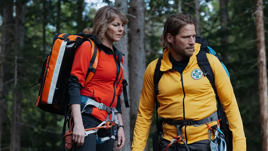 Ausstiege Bergretter Katharina und Markus stehen mit Rucksäcken und Kletterausrüstung im Wald. Sie trägt eine rote Jacke, er eine gelbe.