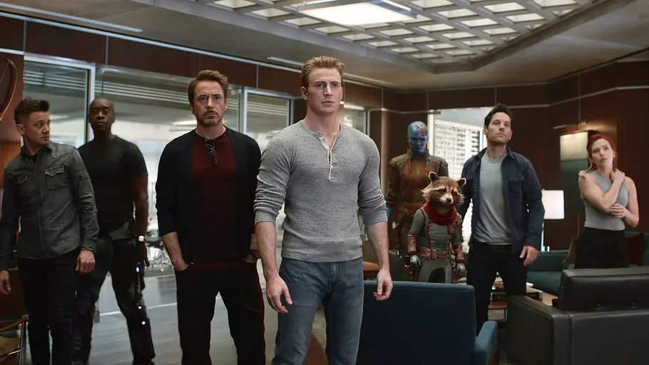 Mehrere Superhelden stehen in einem modern eingerichteten Raum und schauen entschlossen nach vorne. In der Mitte steht ein Mann in grauem Shirt (Captain America), umgeben von weiteren Figuren, darunter Iron Man, Black Widow, War Machine, Hawkeye, Ant-Man, Nebula und Rocket, ein Waschbär in einem Raumanzug.