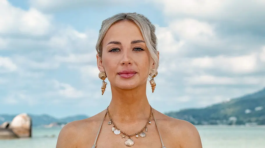 Alexandra Leonhard Eine Frau im Bikini steht am Strand vor dem Meer und blickt ruhig in die Kamera. Sie trägt auffälligen Schmuck und hat die Haare nach hinten gebunden, während im Hintergrund Wasser, Felsen und eine hügelige Küstenlandschaft zu sehen sind.