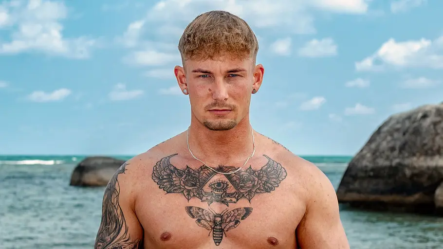 Bennett Bacher Ein tätowierter Mann steht oberkörperfrei am Strand und blickt ernst in die Kamera. Er trägt eine graue Badehose und Schmuck, während im Hintergrund das Meer, große Felsen und ein leicht bewölkter Himmel zu sehen sind.